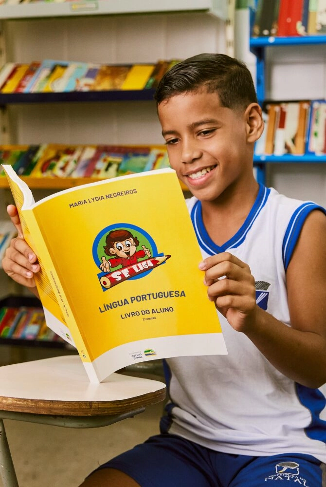 Um menino em uma sala de aula senta-se em uma mesa, sorrindo enquanto lê um livro didático amarelo de língua portuguesa do Plano Anual Instituto Ayrton Senna. As estantes de livros são visíveis ao fundo.