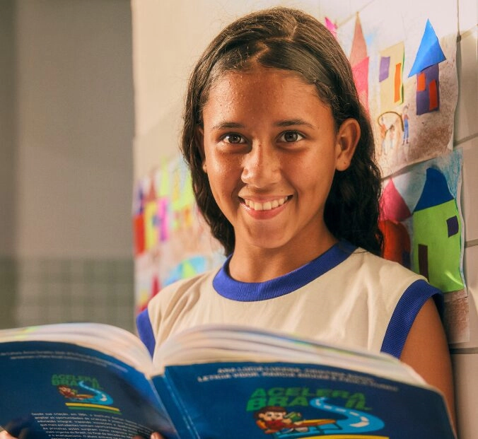 Uma menina em um uniforme escolar sorri enquanto segura um livro aberto, em frente a uma parede decorada com desenhos coloridos de casas.