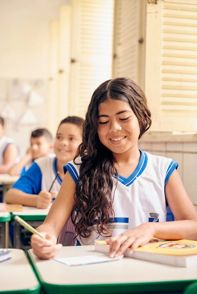 Uma menina em um uniforme escolar sorri enquanto escreve em sua mesa em uma sala de aula, participando de atividades do Plano Anual Instituto Ayrton Senna; outros alunos podem ser vistos ao fundo.