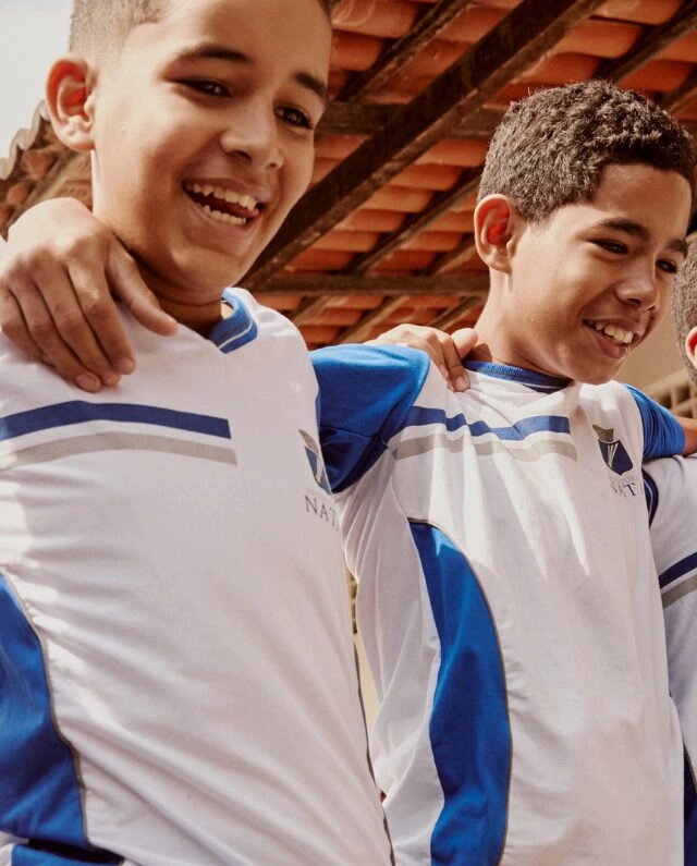Dois meninos vestindo uniformes esportivos brancos e azuis combinando, parte do Plano Anual Instituto Ayrton Senna, estão juntos com os braços em volta dos ombros um do outro, sorrindo.