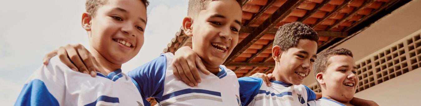 Quatro meninos com uniformes esportivos combinando estão ao ar livre, de braços dados, sorrindo, personificando o espírito de equipe promovido pelo Plano Anual Instituto Ayrton Senna.