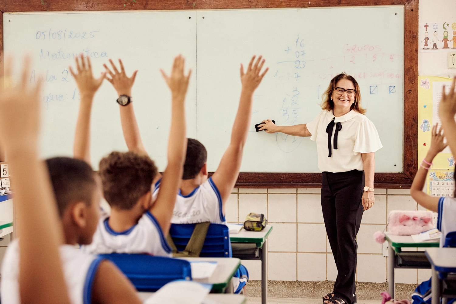 Um professor está ao lado de um quadro branco com problemas de matemática, orientando os alunos que levantam as mãos com entusiasmo em uma sala de aula inspirada no Plano Anual Instituto Ayrton Senna.