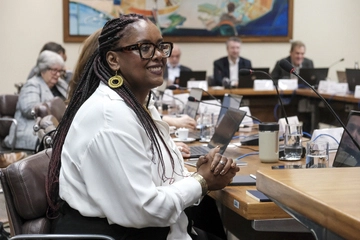 Uma mulher com cabelo trançado e óculos senta-se em uma mesa de conferência do CEE-SP com outras pessoas, cercada por laptops e documentos em uma sala de reuniões.