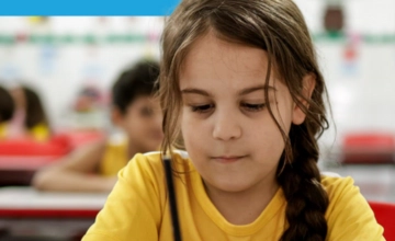 Uma menina de cabelos castanhos trançados, vestindo uma camiseta amarela, senta-se em uma mesa e escreve, com colegas de classe desfocados ao fundo - uma cena do programa Se Liga.