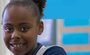 Uma jovem de cabelos cacheados sorri para a câmera em uma sala de aula, com um quadro de avisos azul do Acelera Brasil ao fundo.