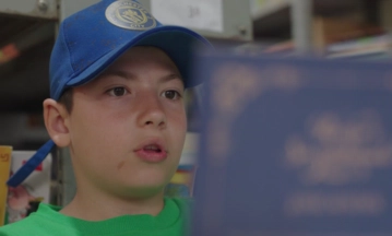 Dia da Poesia - Um garoto usando boné azul e camisa verde olha para um livro azul em uma biblioteca, fazendo anotações como se estivesse escrevendo um rascunho automático.