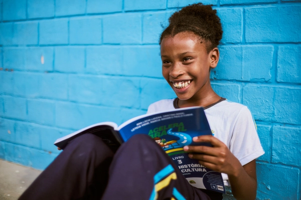 Leitura e Repertório Cultural - uma criança sorridente senta-se contra uma parede azul brilhante, segurando e lendo um livro escolar aberto intitulado "História e Cultura", com anotações e rascunho automático rabiscados nas margens.