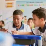 Dois meninos em uniformes escolares trabalham juntos em uma mesa em uma sala de aula, refletindo o espírito colaborativo promovido pelo Plano Anual Instituto Ayrton Senna, com outros alunos e cartazes educacionais visíveis ao fundo.