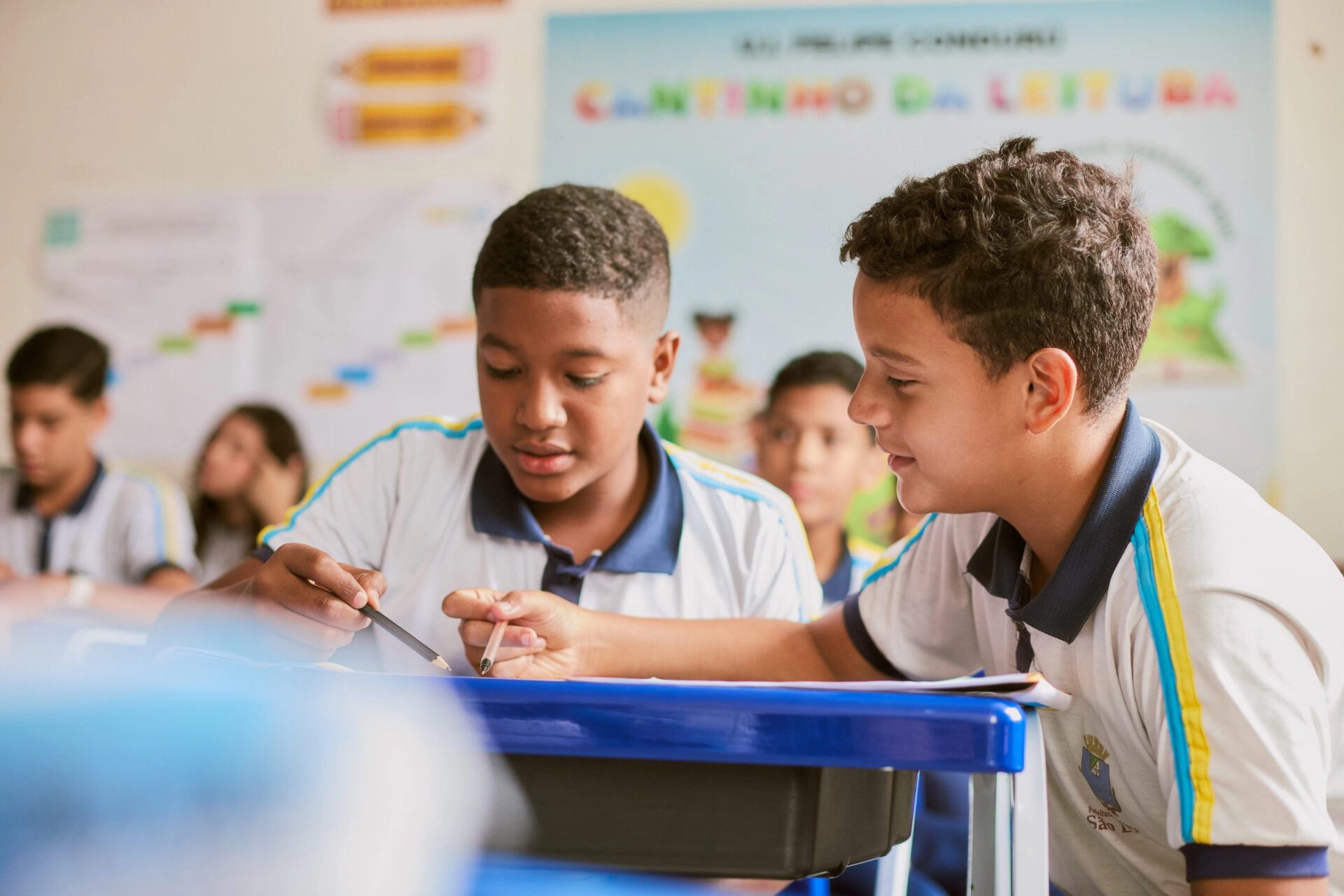 Dois meninos em uniformes escolares trabalham juntos em uma mesa em uma sala de aula, refletindo o espírito colaborativo promovido pelo Plano Anual Instituto Ayrton Senna, com outros alunos e cartazes educacionais visíveis ao fundo.
