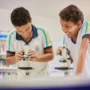Dois alunos usando uniformes escolares examinam amostras com microscópios em uma sala de aula ou biblioteca. As estantes de livros são visíveis ao fundo.
