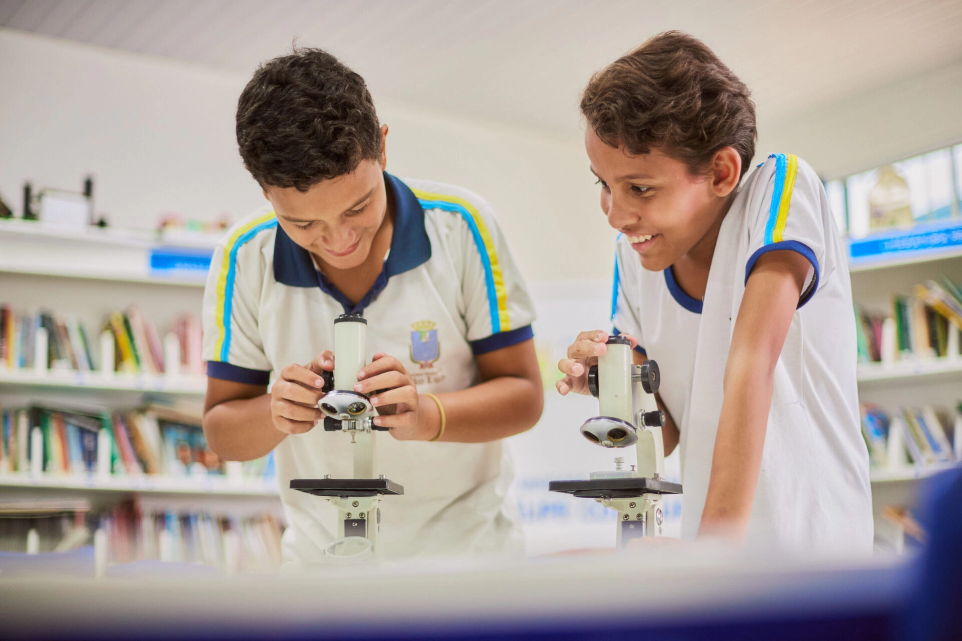 Dois alunos usando uniformes escolares examinam amostras com microscópios em uma sala de aula ou biblioteca. As estantes de livros são visíveis ao fundo.