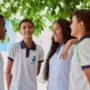 Quatro alunos com uniformes iguais ficam de pé e conversam ao ar livre sob uma árvore.