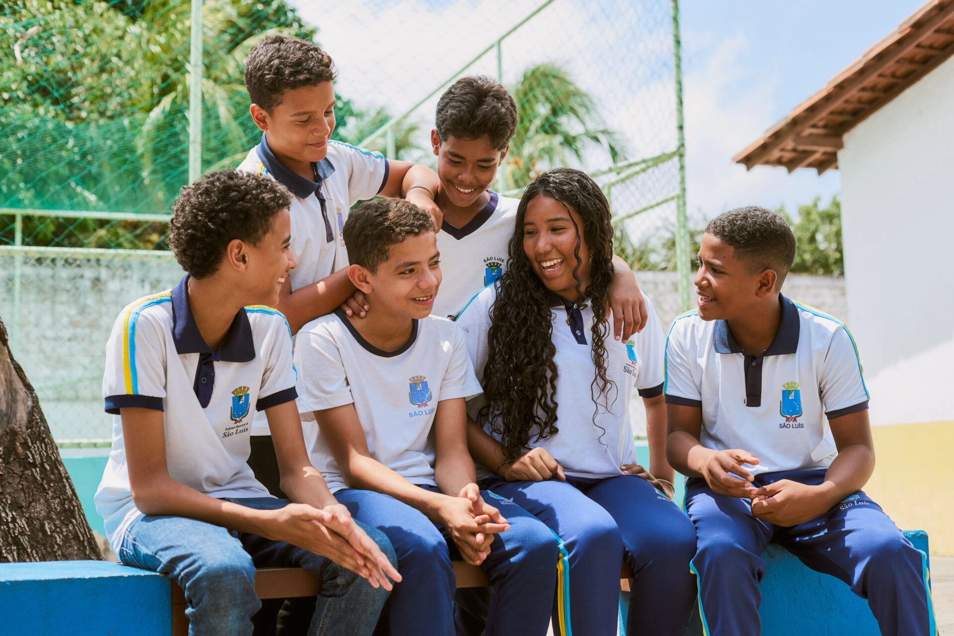 Seis alunos com uniformes iguais sentam-se e ficam juntos em um banco ao ar livre, sorrindo e conversando perto de uma quadra de esportes.