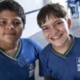 Dois meninos com uniformes escolares azuis estão em pé dentro de casa, olhando para a câmera. Um deles sorri amplamente, enquanto o outro tem uma expressão neutra. Outros alunos podem ser vistos ao fundo.