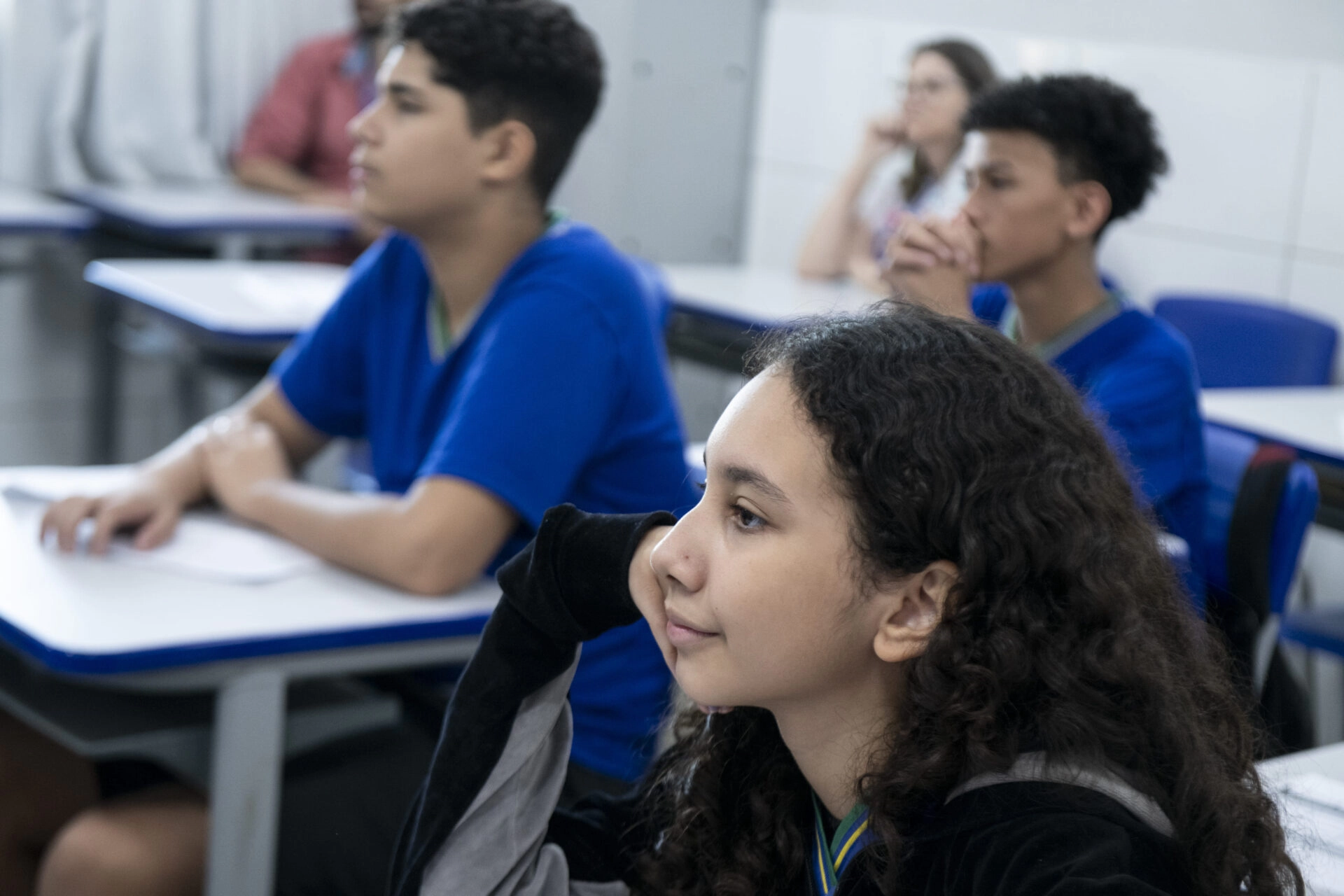 Os alunos sentam-se em carteiras em uma sala de aula, ouvindo atentamente e olhando para a frente da sala.