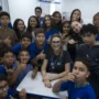Um professor senta-se em uma mesa cercado por alunos sorridentes em uma sala de aula, a maioria deles usando uniformes azuis e fazendo gestos com o polegar para cima.