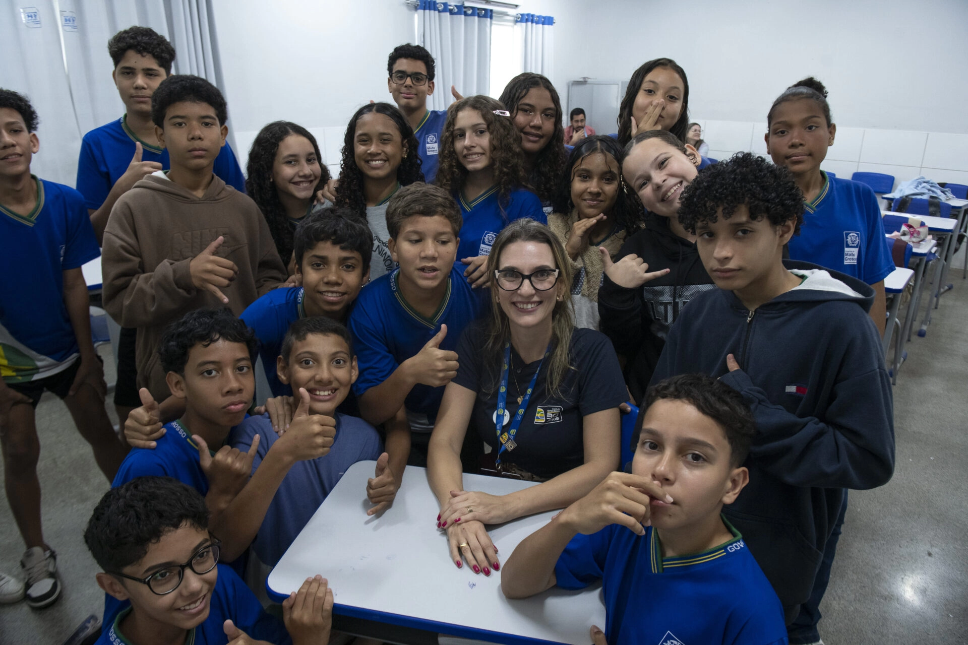 Um professor senta-se em uma mesa cercado por alunos sorridentes em uma sala de aula, a maioria deles usando uniformes azuis e fazendo gestos com o polegar para cima.