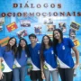 Cinco alunos com uniformes azuis e brancos estão sorridentes em frente a uma parede azul decorada com fotos e os dizeres "Diálogos Socioemocionais".