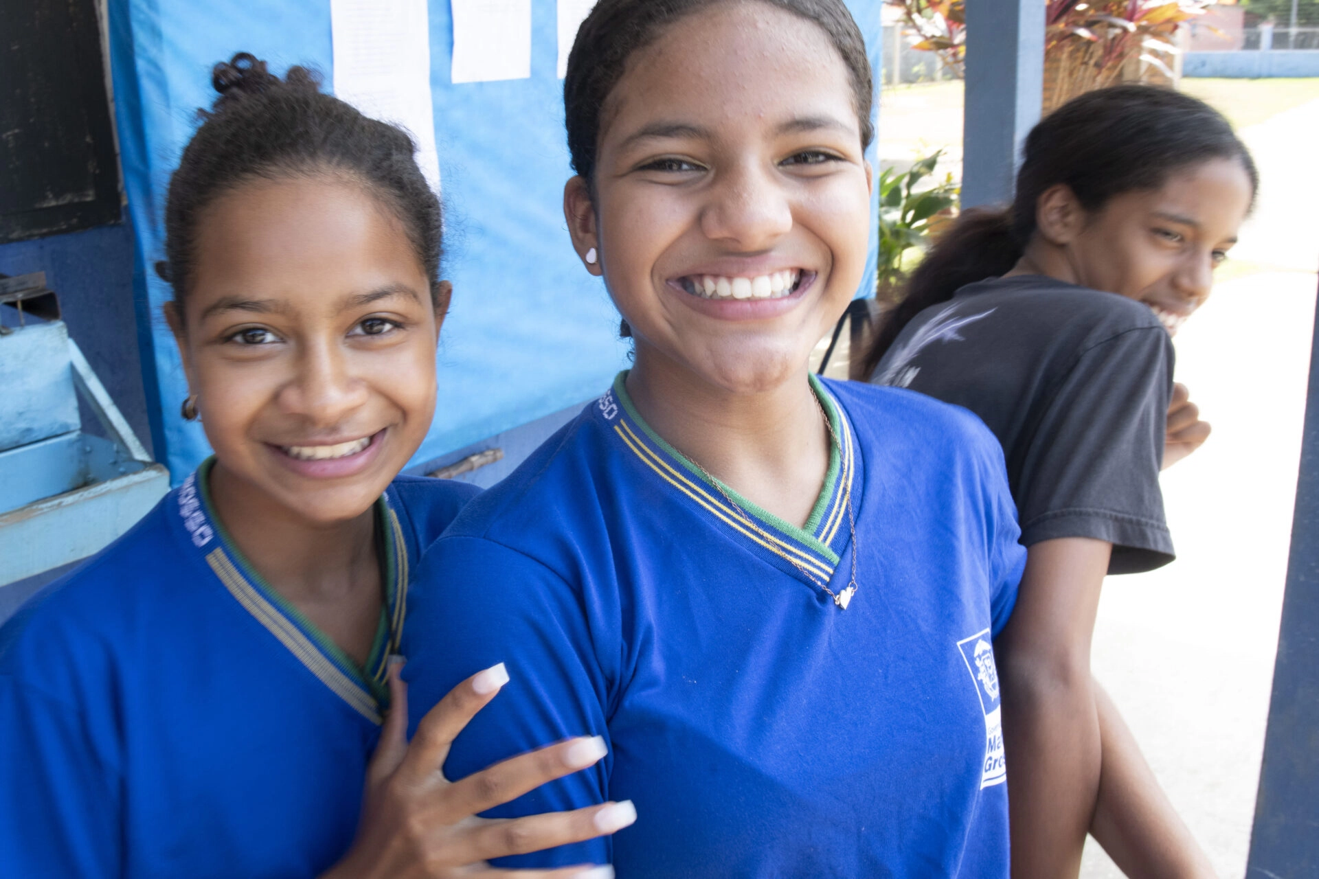Três alunos estão juntos, dois sorrindo para a câmera com uniformes azuis, enquanto um de camisa preta olha para o fundo.