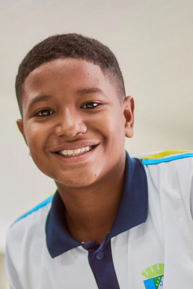 Um menino de cabelos curtos sorri para a câmera, vestindo uma camisa branca com gola azul e detalhes em verde e azul no ombro e no peito, representando orgulhosamente o Projeto Apoio Cultural Instituto Ayrton Senna.