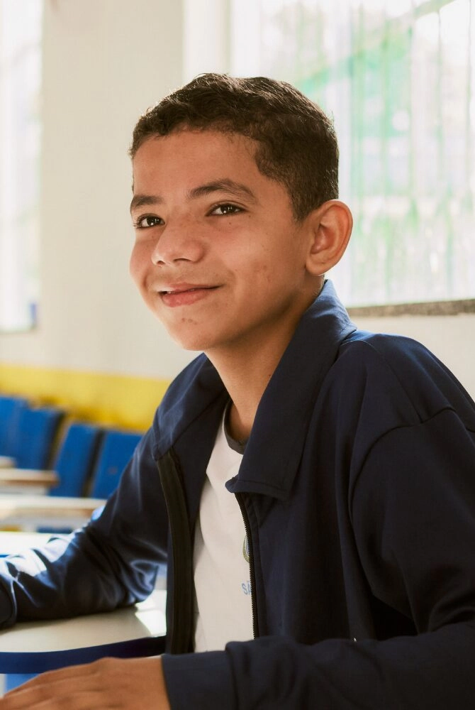 Um menino de cabelos curtos e cacheados senta-se em uma mesa em uma sala de aula, vestindo uma jaqueta escura e camisa branca, sorrindo levemente para a câmera como parte do Projeto Apoio Cultural Instituto Ayrton Senna.