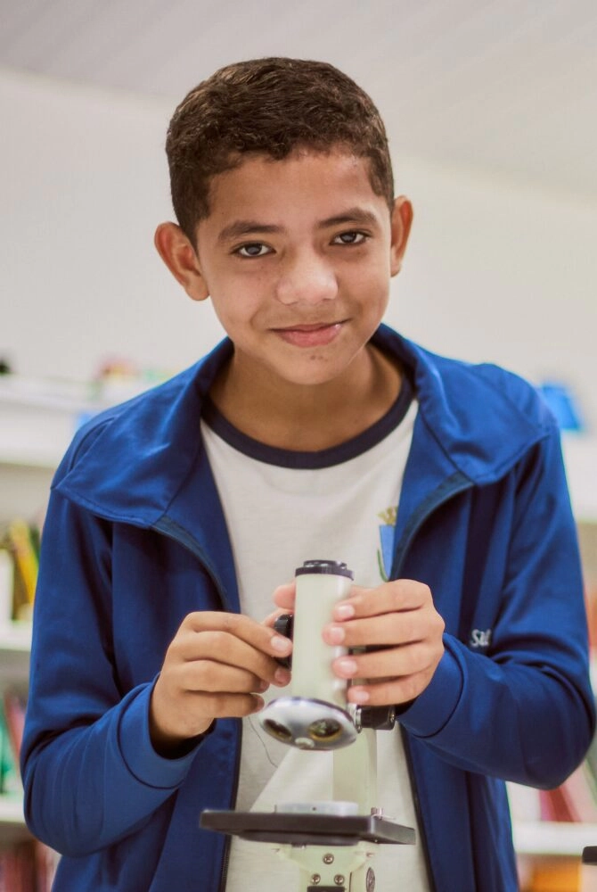Um menino vestindo uma jaqueta azul está em pé dentro de casa, sorrindo e segurando um microscópio, com estantes de livros visíveis ao fundo - um momento inspirador alinhado com o compromisso do Plano Anual Instituto Ayrton Senna com a educação.