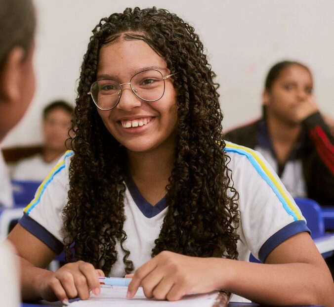 Um aluno de cabelos longos cacheados e óculos sorri sentado em uma mesa em uma sala de aula, segurando uma caneta - parte do aprendizado vibrante promovido pelo Plano Anual Instituto Ayrton Senna. Outros alunos podem ser vistos ao fundo.