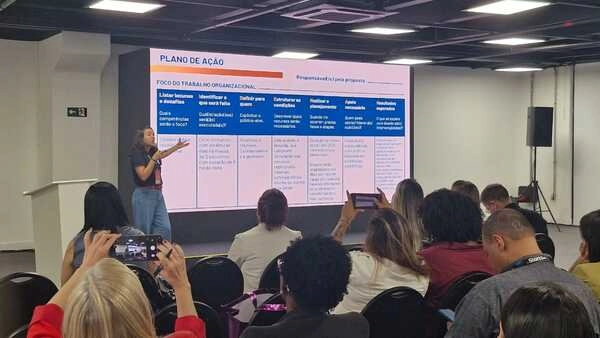 Uma mulher apresenta um slide de plano de ação para um público em uma sala de conferências no RH Leadership Festival; os participantes ouvem e tiram fotos.
