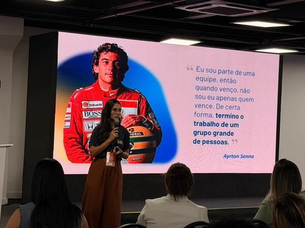 Uma mulher se apresenta diante de uma plateia no RH Leadership Festival, com um telão mostrando Ayrton Senna em um equipamento de corrida e uma citação em português atribuída a ele.
