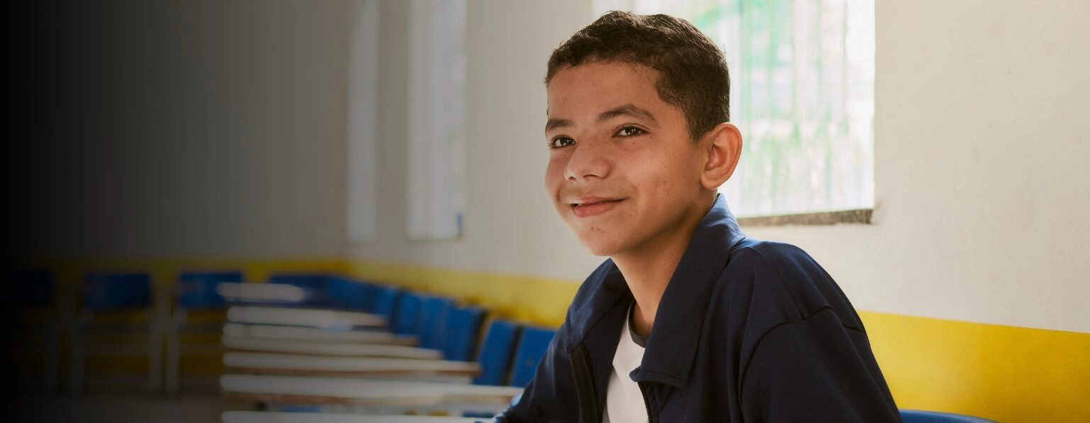 Um menino vestindo uma jaqueta escura senta-se em uma mesa em uma sala de aula com cadeiras azuis e acabamento amarelo, sorrindo para a câmera, representando orgulhosamente o Instituto Ayrton Senna.