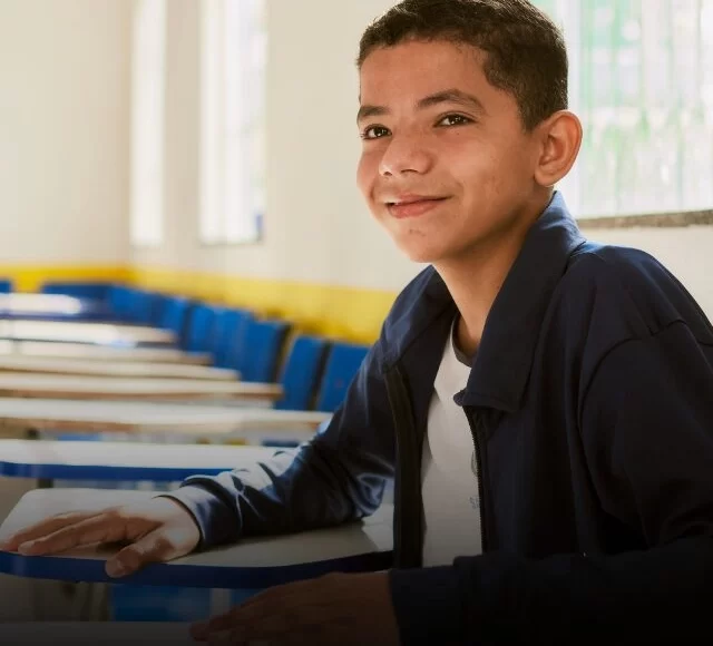 Um menino vestindo uma jaqueta escura senta-se em uma mesa em uma sala de aula com cadeiras azuis e acabamento amarelo, sorrindo para a câmera, representando orgulhosamente o Instituto Ayrton Senna.