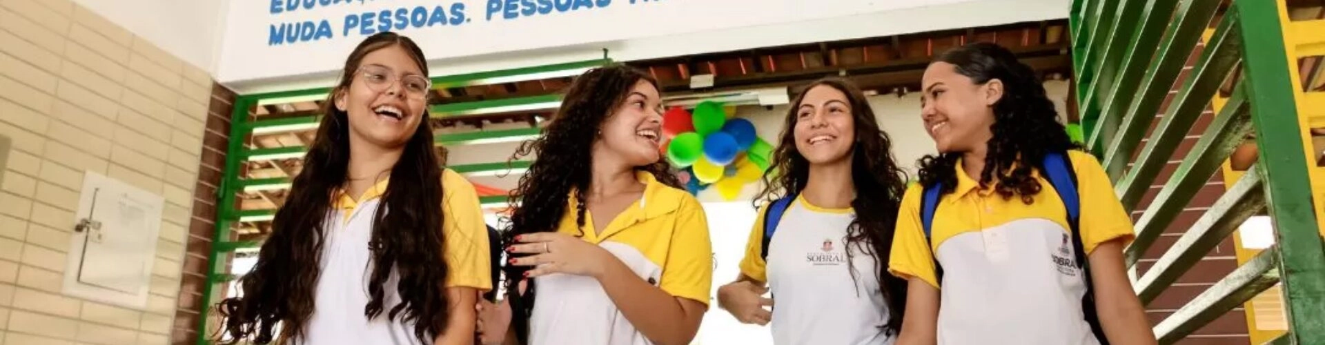 Quatro alunas com uniformes escolares caminham juntas em um corredor, sorrindo e conversando. Balões coloridos e uma placa motivacional são visíveis ao fundo, refletindo temas de uma coluna recente da Revista Educação.