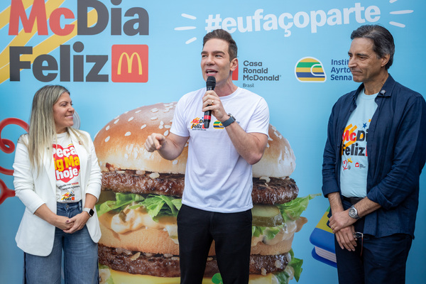 Três pessoas estão em frente a um cenário do evento “McDia Feliz 2026”, com a imagem de um hambúrguer; uma delas fala ao microfone sobre oportunidades e a importância da educação pública, enquanto as outras ouvem atentamente.
