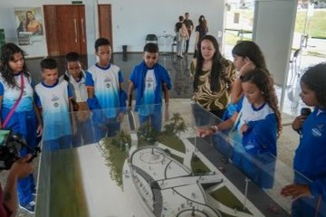 Um grupo de crianças e um adulto, que lembram os estudantes que visitaram o Teatro Amazonas, observam uma maquete arquitetônica exposta sobre uma mesa de vidro dentro de um prédio moderno.