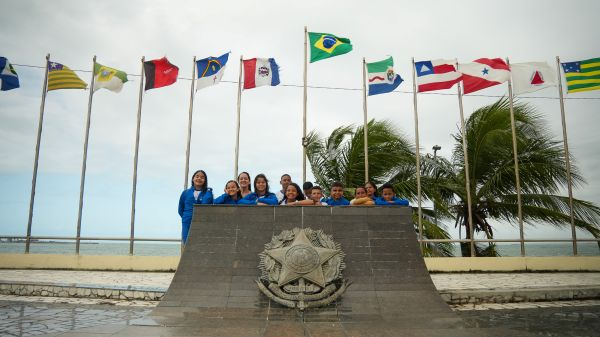 Um grupo de pessoas vestindo uniformes azuis está em pé atrás de um monumento no Memorial à República, tendo ao fundo várias bandeiras estaduais e palmeiras sob um céu nublado.