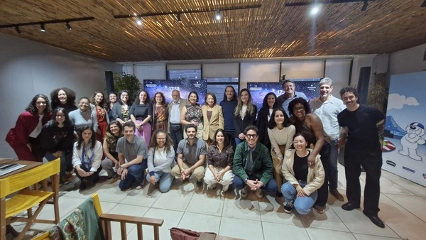 Um grande grupo de pessoas posando para uma foto em um evento do Instituto Ayrton Senna, com telas de avaliação educacional ao fundo.