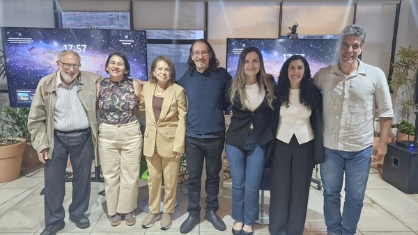 Sete pessoas posam para uma foto em grupo em frente a monitores que exibem imagens espaciais, durante um evento de avaliação educacional do Instituto Ayrton Senna.