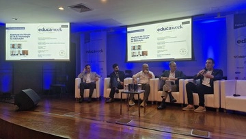 Cinco pessoas estão sentadas em poltronas brancas no palco da Educa Week 2026, com duas telas exibindo a programação do evento atrás delas.