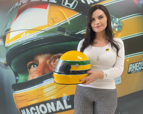 Uma mulher segura um capacete de corrida amarelo em frente a uma fotografia de Ayrton Senna que ocupa toda a parede na Cátedra Instituto Ayrton Senna.